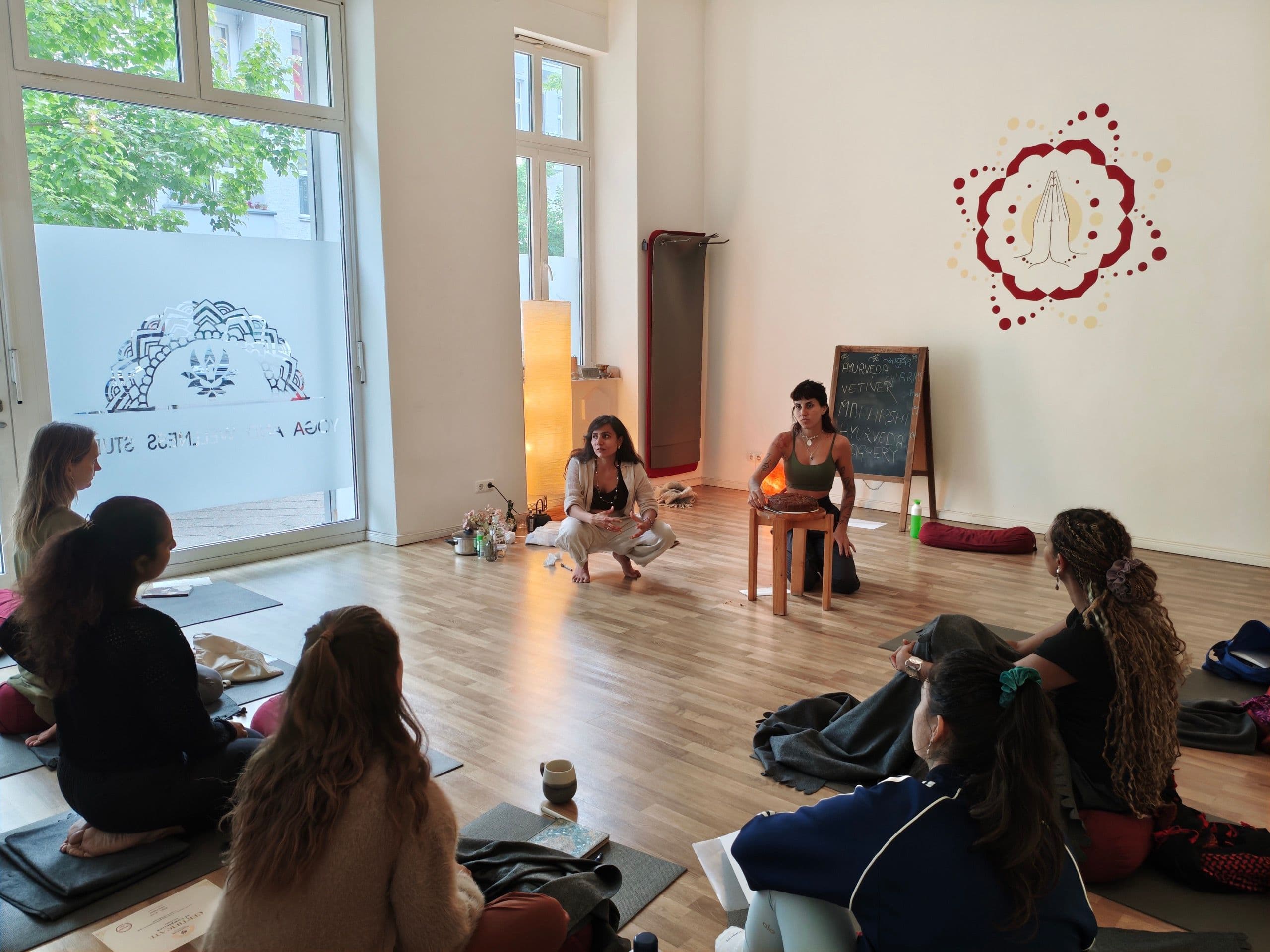 Students sit in a circle on mats while two teachers speak at the front of a bright studio