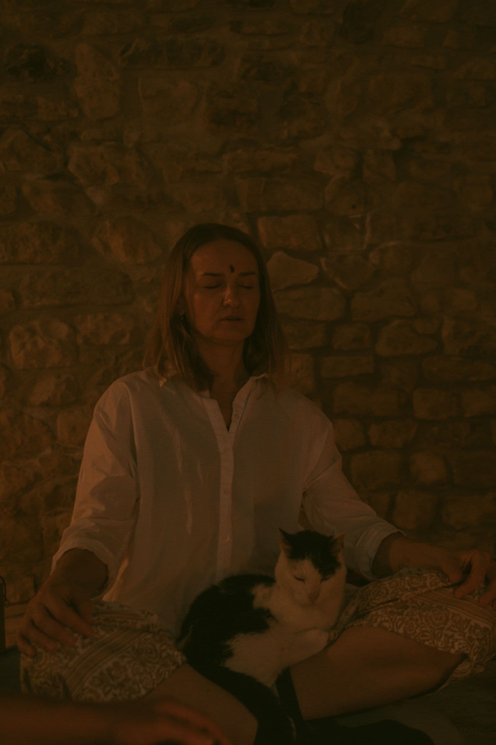 A woman meditates in candlelight against a stone wall with a cat asleep in her lap