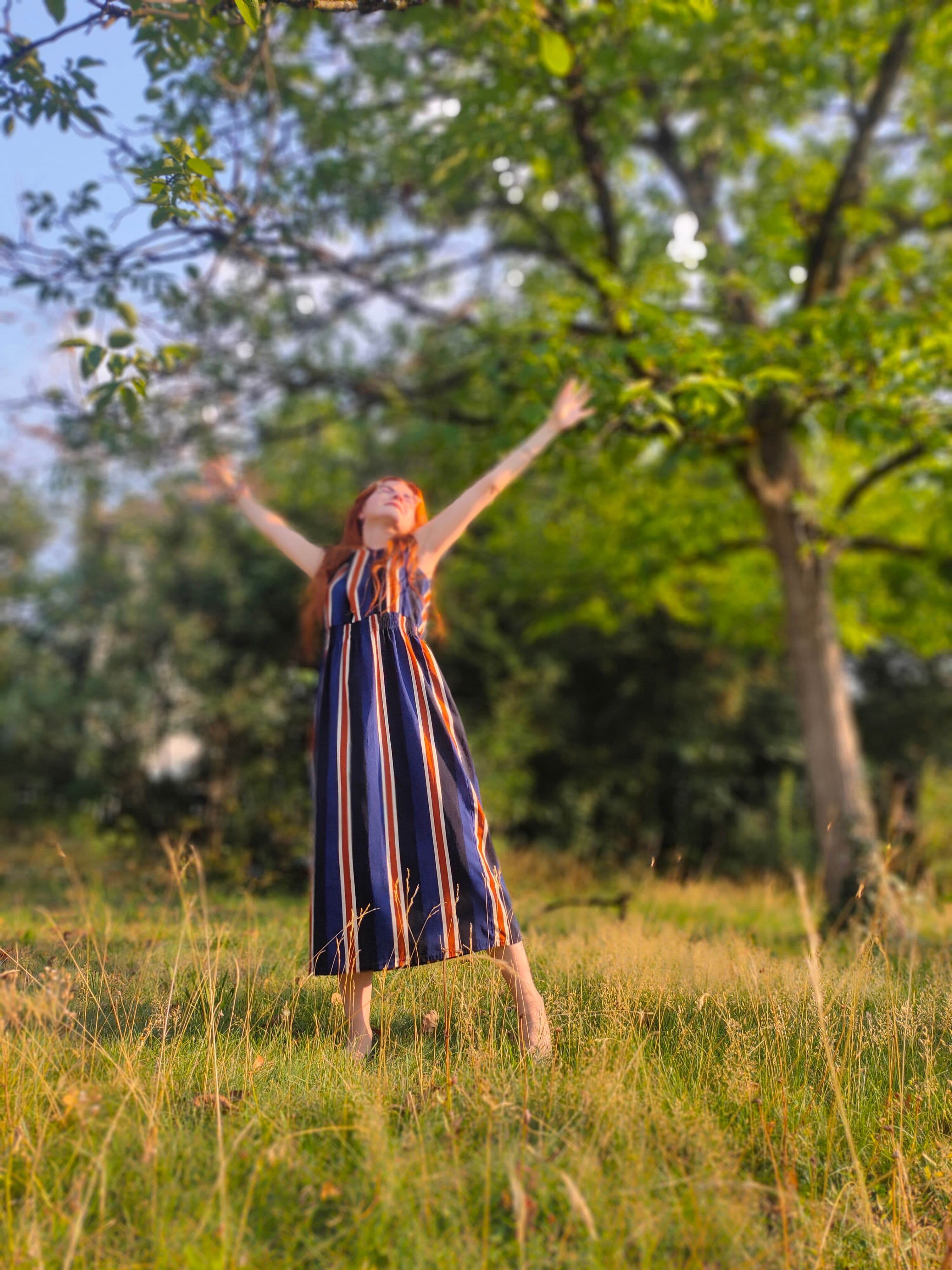 Luisa practicing a joyful yoga-inspired standing stretch outdoors, barefoot in a sunlit meadow with her arms raised toward the sky, surrounded by lush green trees and nature.