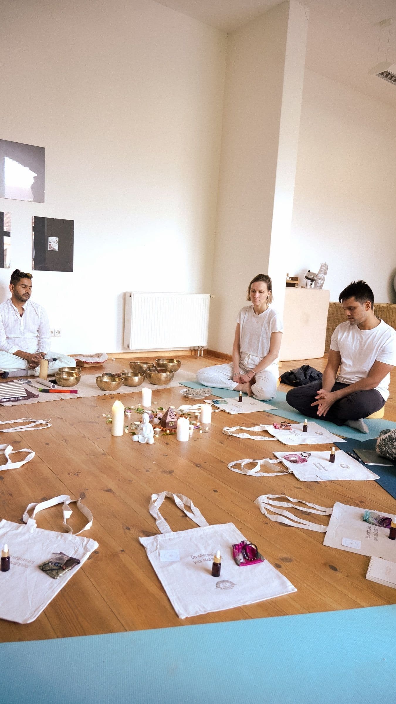 Three people in white sit in a circle on a wooden studio floor around singing bowls, candles and tote bags